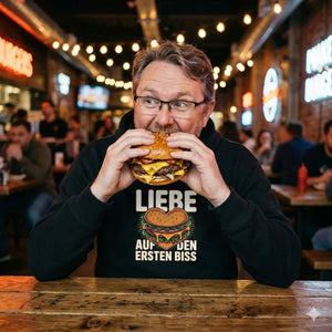 Ein Mann mit Brille beißt genussvoll in einen großen Burger, trägt einen schwarzen Hoodie mit dem Aufdruck "LIEBE AUF DEN ERSTEN BISS" in einem warm beleuchteten Restaurant.