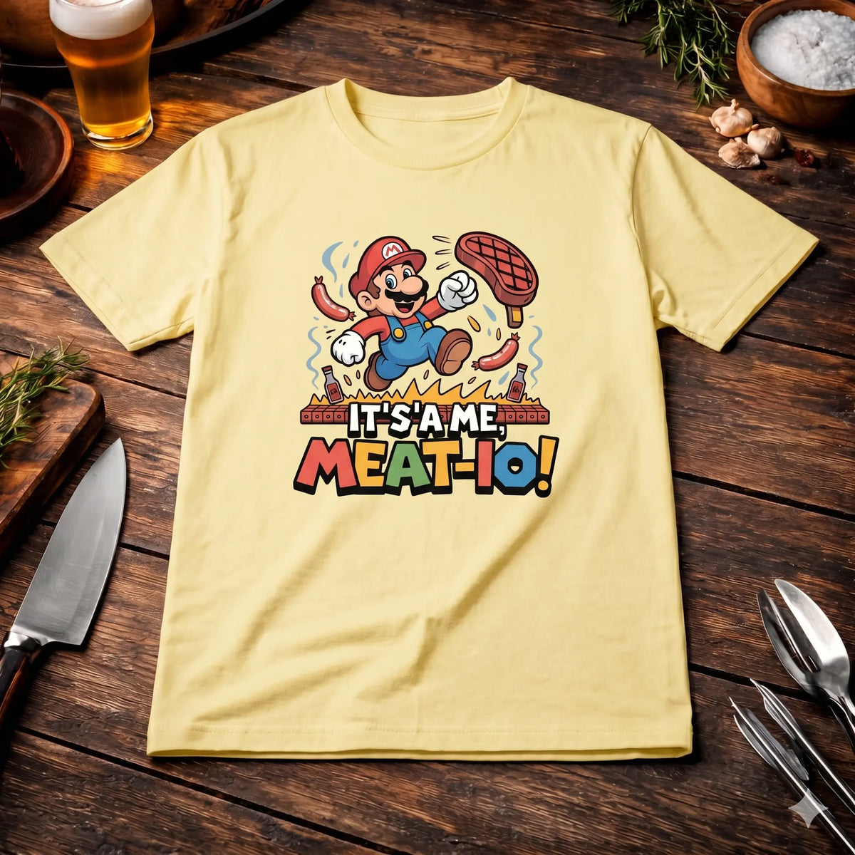 Ein hellgelbes T-Shirt mit einem cartoonartigen Aufdruck von Mario, der über gegrillten Fleischstücken und Hot Dogs springt. Der Text „IT'S A ME, MEAT-IO!“ ist darunter in großen, farbigen Buchstaben zu lesen. Das T-Shirt liegt flach auf einer dunklen Holzoberfläche, umgeben von einem Messer, Gabeln, Grillspießen, einem Glas Bier, frischen Rosmarinzweigen und Schalen mit Knoblauch und Meersalz. Das Bild ist aus der Vogelperspektive aufgenommen.