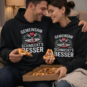 Glückliches Paar auf der Couch teilt Pizza und trägt schwarze Meatware Hoodies mit dem Spruch „Gemeinsam schmeckt’s besser“ – Liebe geht durch den Magen, gemütlicher Abendlook.