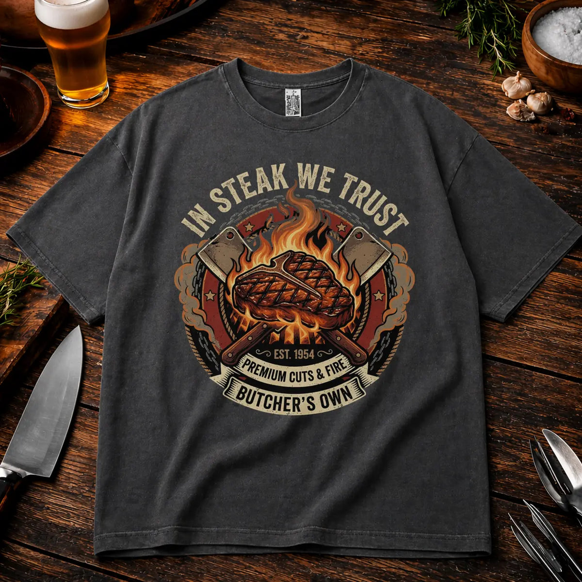 Graphitfarbenes Oversized T-Shirt mit „In Steak We Trust“ Grillmotiv im rustikalen BBQ Mockup auf Holztisch mit Bier, Messer und Gewürzen, lockerer Schnitt, realistische Stoffstruktur und hochwertiger Druck.