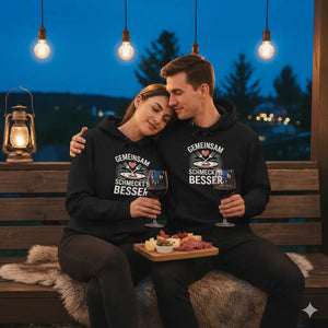 Paar kuschelt auf Holzbank bei Laternenschein, trägt schwarzen Premium Hoodie mit „Gemeinsam schmeckt’s besser“ Design.