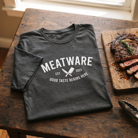 Gefaltetes anthrazitfarbenes Meatware-Shirt mit weißem Aufdruck neben gegrilltem Steak und Rosmarin auf einem rustikalen Holztisch.