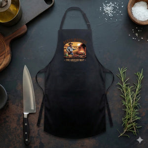 Ein professionelles Mockup einer schwarzen Grillschürze, flach liegend auf einer rustikalen, dunklen Arbeitsplatte. Die Schürze ist mit dem „Asado Masters - The Gaucho Way“-Logo bedruckt. Die Szene wird durch kulinarische Accessoires ergänzt: Ein scharfes Küchenmesser, frischer Rosmarin, eine Schale mit Meersalz und eine Flasche Öl unterstreichen den hochwertigen Grill- und Koch-Kontext.