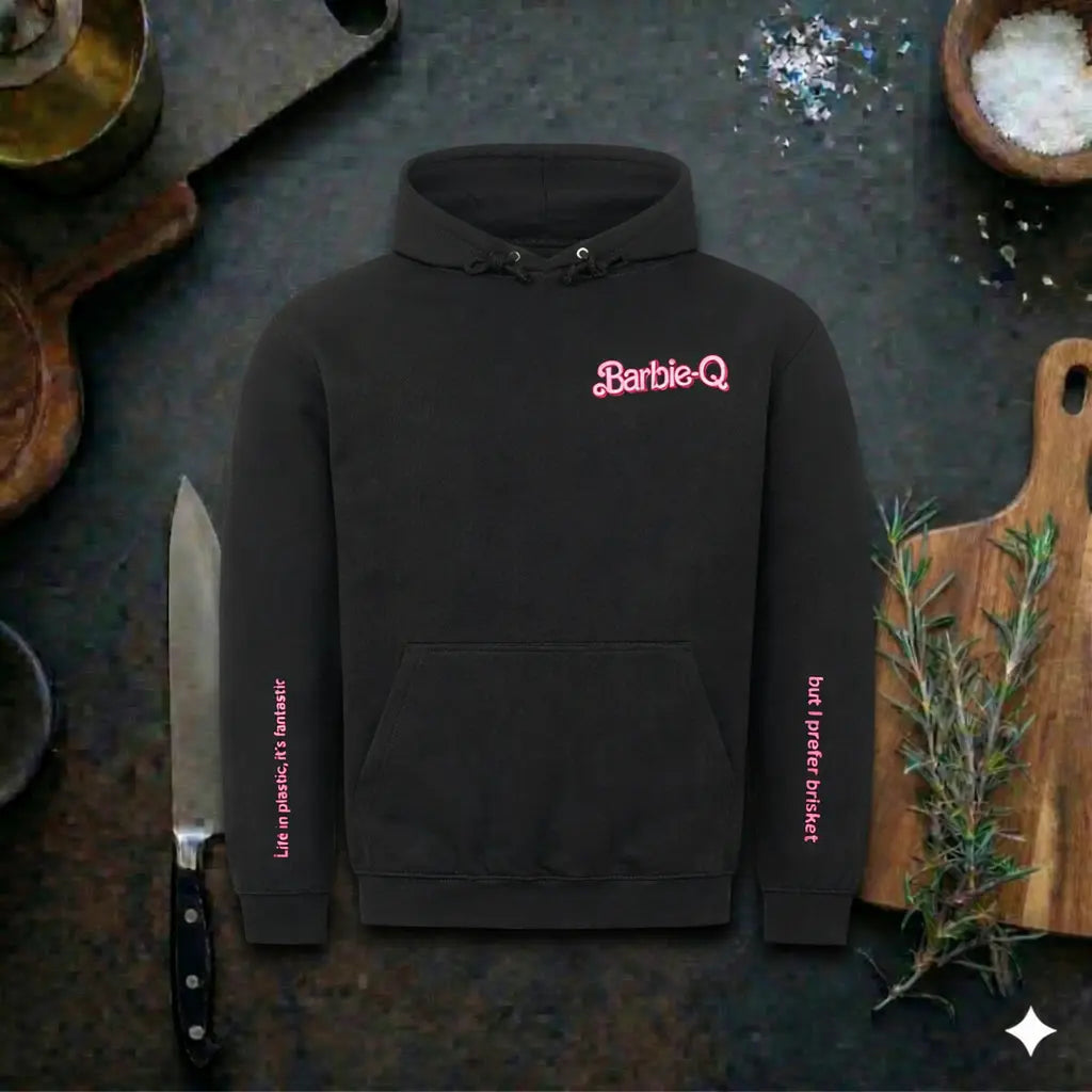 Black BBQ hoodie with pink Barbie-Q logo and sleeve text, Meatware grill hoodie displayed in rustic kitchen mockup with knife, cutting board and herbs