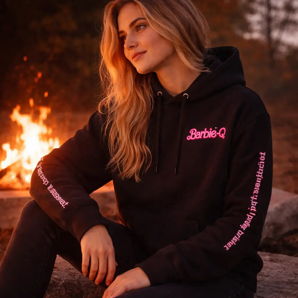 Frau trägt schwarzen Barbie-Q BBQ Hoodie mit pinkem Schriftzug und Ärmeldruck am Lagerfeuer, Grill Lifestyle Hoodie mit Spruch „Life in plastic it's fantastic but I prefer brisket“