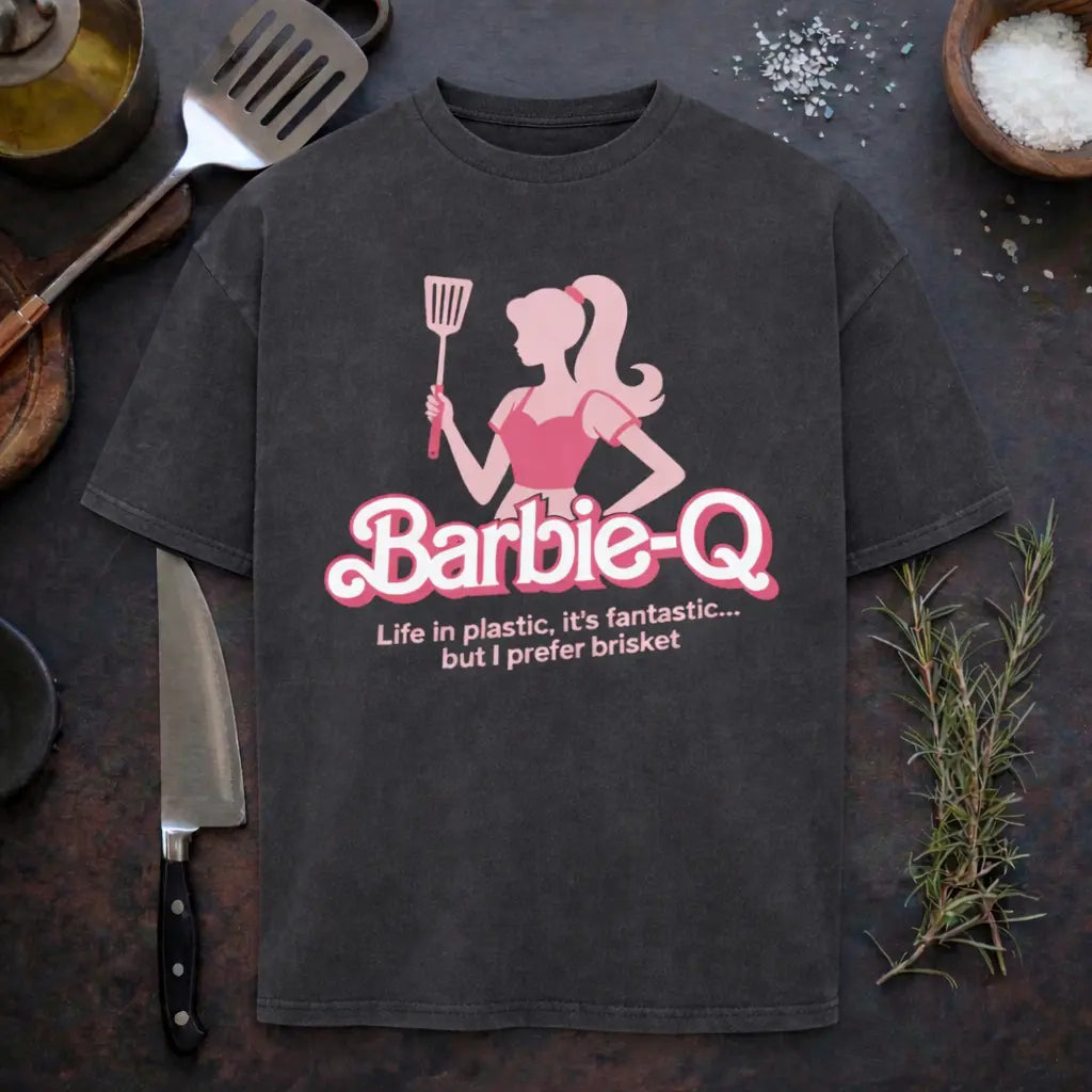 Oversized BBQ Humor T-Shirt „Barbie-Q“ mit pinker Grill-Silhouette und Spruch „Life in plastic, it’s fantastic… but I prefer brisket“, präsentiert in rustikalem BBQ-Küchen-Mockup mit Grillspatel, Kochmesser, Rosmarin und grobem Salz.