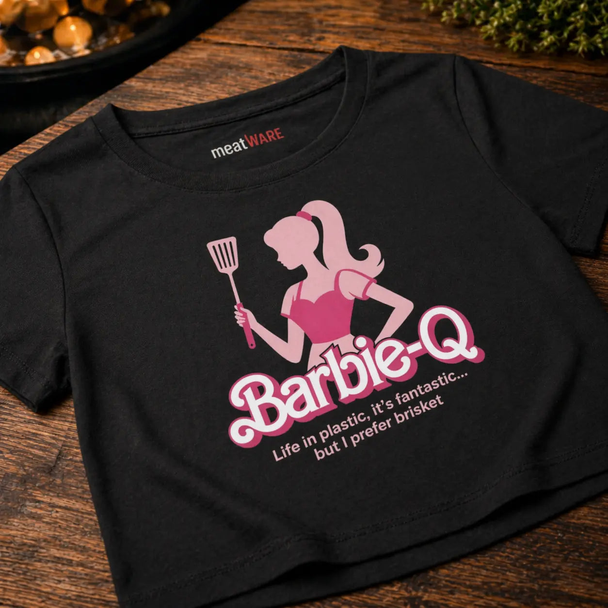 Eine junge Frau in einem schwarzen Barbie-Q Crop Top steht lächelnd hinter einem schwarzen Kugelgrill und hält eine Grillzange über der Schulter. Im sonnigen Hintergrund ist eine pinke Luftmatratze zu sehen.