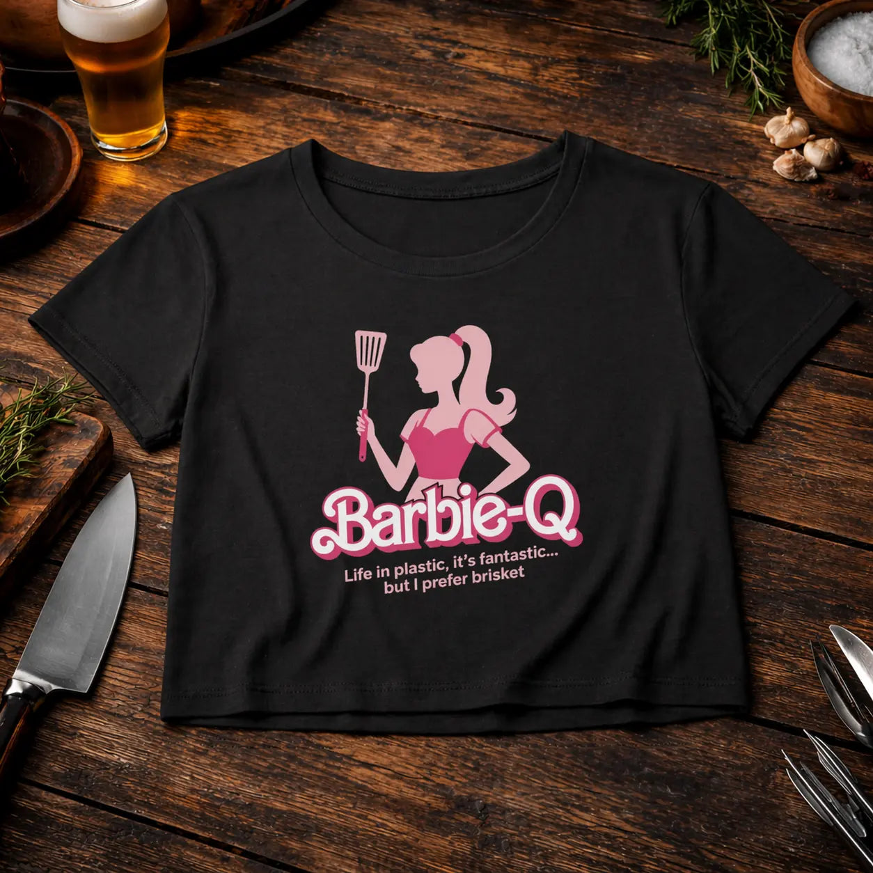 Black ladies cropped tee with Barbie-Q brisket BBQ design in pink, styled on rustic wooden table with beer, grill tools and herbs – Meatware Shirts & more mit Geschmack