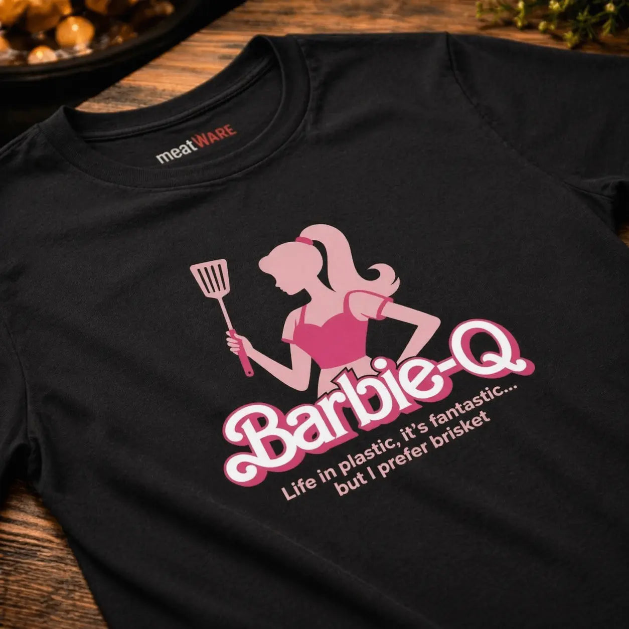 Close-up of premium organic women T-shirt with Barbie-Q brisket BBQ design in pink on black fabric, detailed print view on rustic wooden table – Meatware Shirts & more mit Geschmack