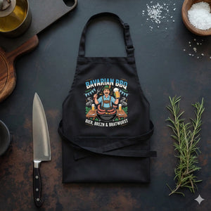 Eine schwarze Latzschürze auf einer dunklen Küchenarbeitsplatte, platziert zwischen einem Kochmesser und frischem Rosmarin. Auf der Schürze ist ein farbenfrohes Design mit einem Mann in bayerischer Tracht beim Grillen sowie dem Text „Bavarian BBQ – Bier, Brezn & Bratwurst“ zu sehen.