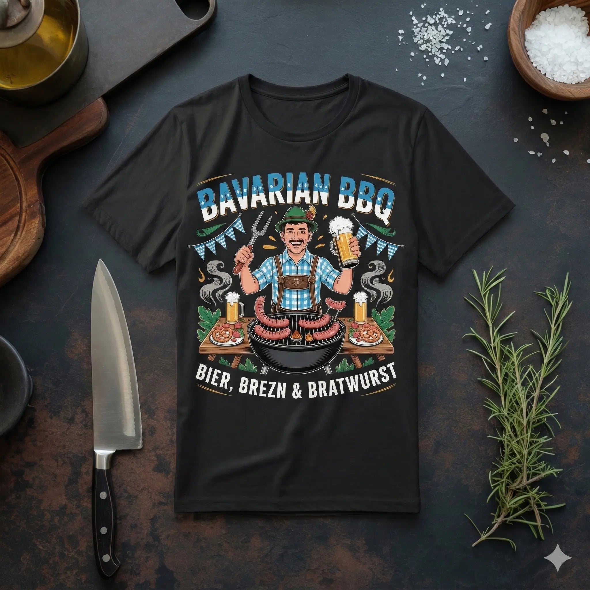 Ein schwarzes T-Shirt auf einer dunklen Arbeitsplatte neben einem Küchenmesser und Rosmarin. Das Design zeigt einen lächelnden Mann in Lederhosen beim Grillen mit Bier, Brezen und Bratwurst unter dem Schriftzug „Bavarian BBQ“.