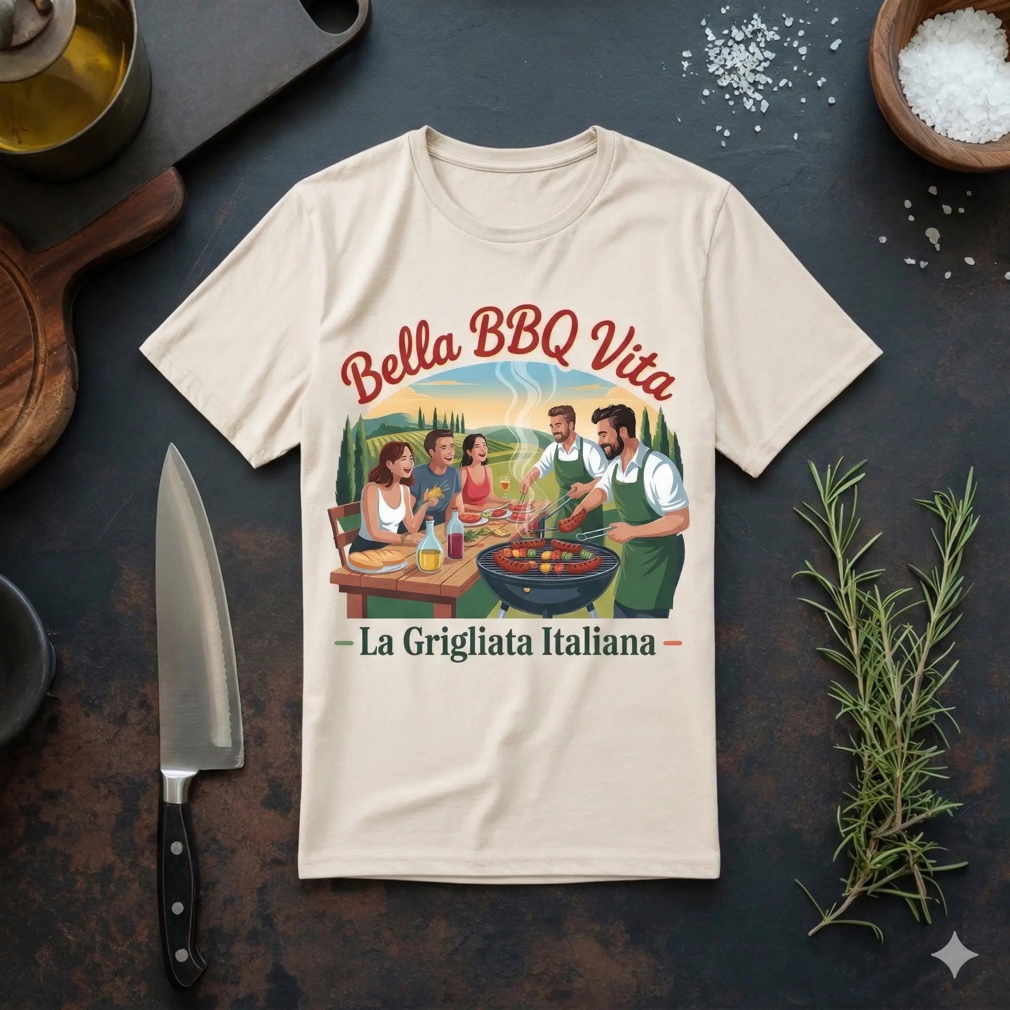 Ein hellnaturfarbenes (cremefarbenes) Heavy Cotton Oversized T-Shirt mit dem Aufdruck ‚Bella BBQ Vita – La Grigliata Italiana‘ liegt flach auf einer dunklen Schiefer-Arbeitsplatte. Das Design zeigt eine Gruppe von fünf Freunden, die an einem Tisch im Freien sitzen und grillen, mit einer toskanischen Landschaft im Hintergrund. Um das T-Shirt herum sind Küchenutensilien wie ein großes Kochmesser, ein Holzschneidebrett, Ölkännchen, Schalen mit Meersalz und frischer Rosmarin angeordnet