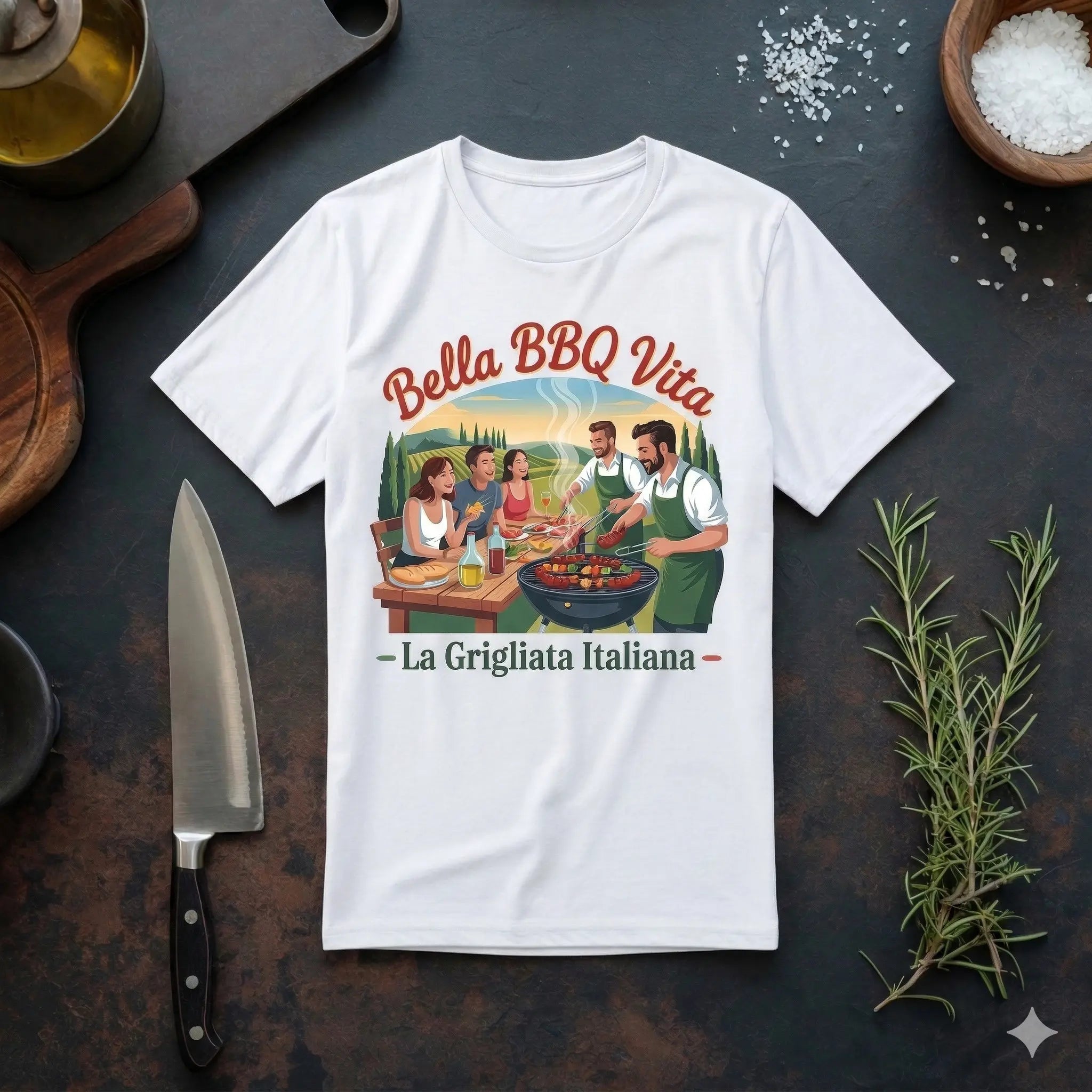 Ein weißes T-Shirt mit dem Aufdruck ‚Bella BBQ Vita – La Grigliata Italiana‘ liegt flach auf einer dunklen Schiefer-Arbeitsplatte. Das Design zeigt eine Gruppe von fünf Freunden, die an einem Tisch im Freien sitzen und grillen, mit einer toskanischen Landschaft im Hintergrund. Um das T-Shirt herum sind Küchenutensilien wie ein großes Kochmesser, ein Holzschneidebrett, Ölkännchen, Schalen mit Meersalz und frischer Rosmarin angeordnet.