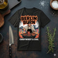 Ein schwarzes T-Shirt mit einem urbanen Grill-Design flach liegend auf einer dunklen Arbeitsplatte. Das Motiv zeigt einen Mann am Grill vor einer Berliner Skyline-Kulisse mit den Schriftzügen „BERLIN BURN“ und „CURRYWURST & COAL“. Daneben liegen ein Messer und Rosmarin