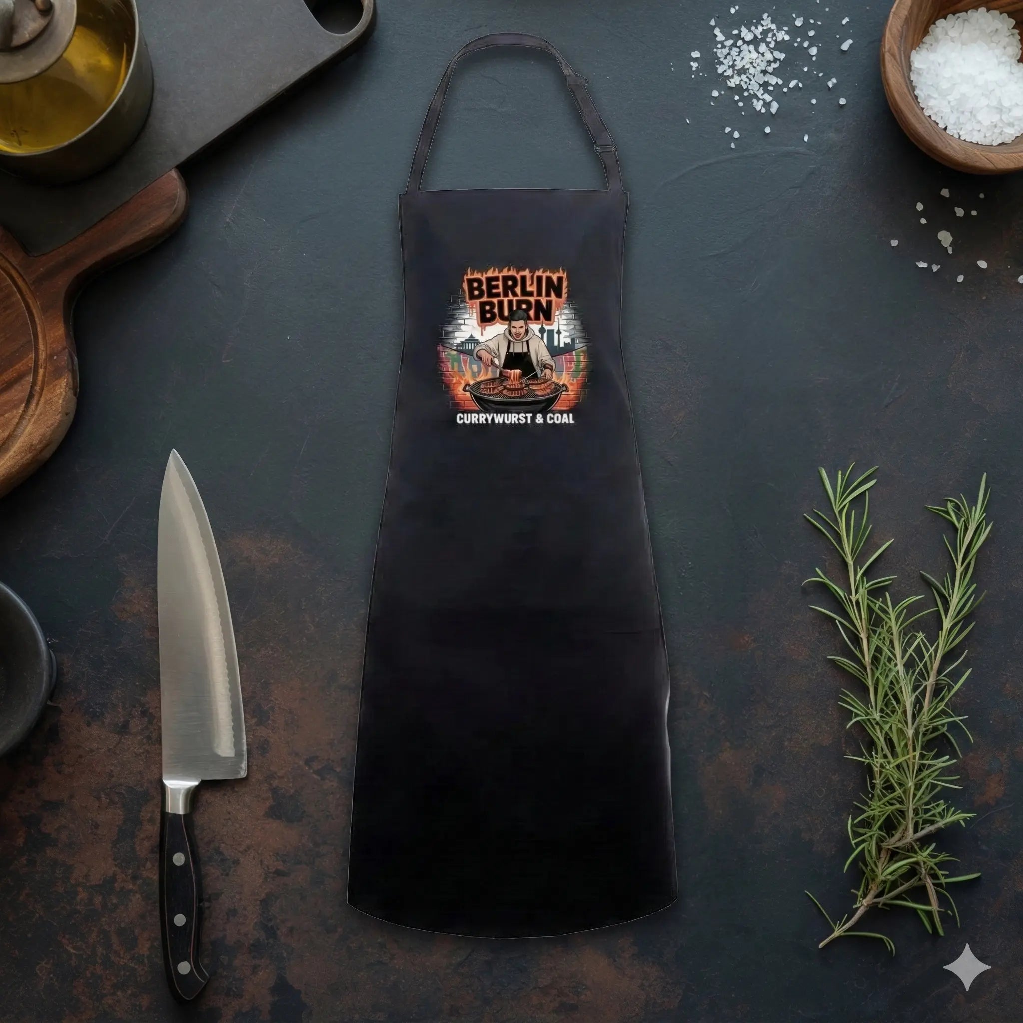 „Eine schwarze Grillschürze mit dem Aufdruck ‚Berlin Burn – Currywurst & Coal‘ liegt flach auf einer dunklen Arbeitsplatte aus Schiefer. Der Aufdruck zeigt einen Cartoon-Koch, der vor der Berliner Skyline grillt. Um die Schürze herum sind Küchenwerkzeuge wie ein großes Kochmesser, ein Holzschneidebrett, ein Ölkännchen, Schalen mit Meersalz und ein Zweig frischer Rosmarin angeordnet.“