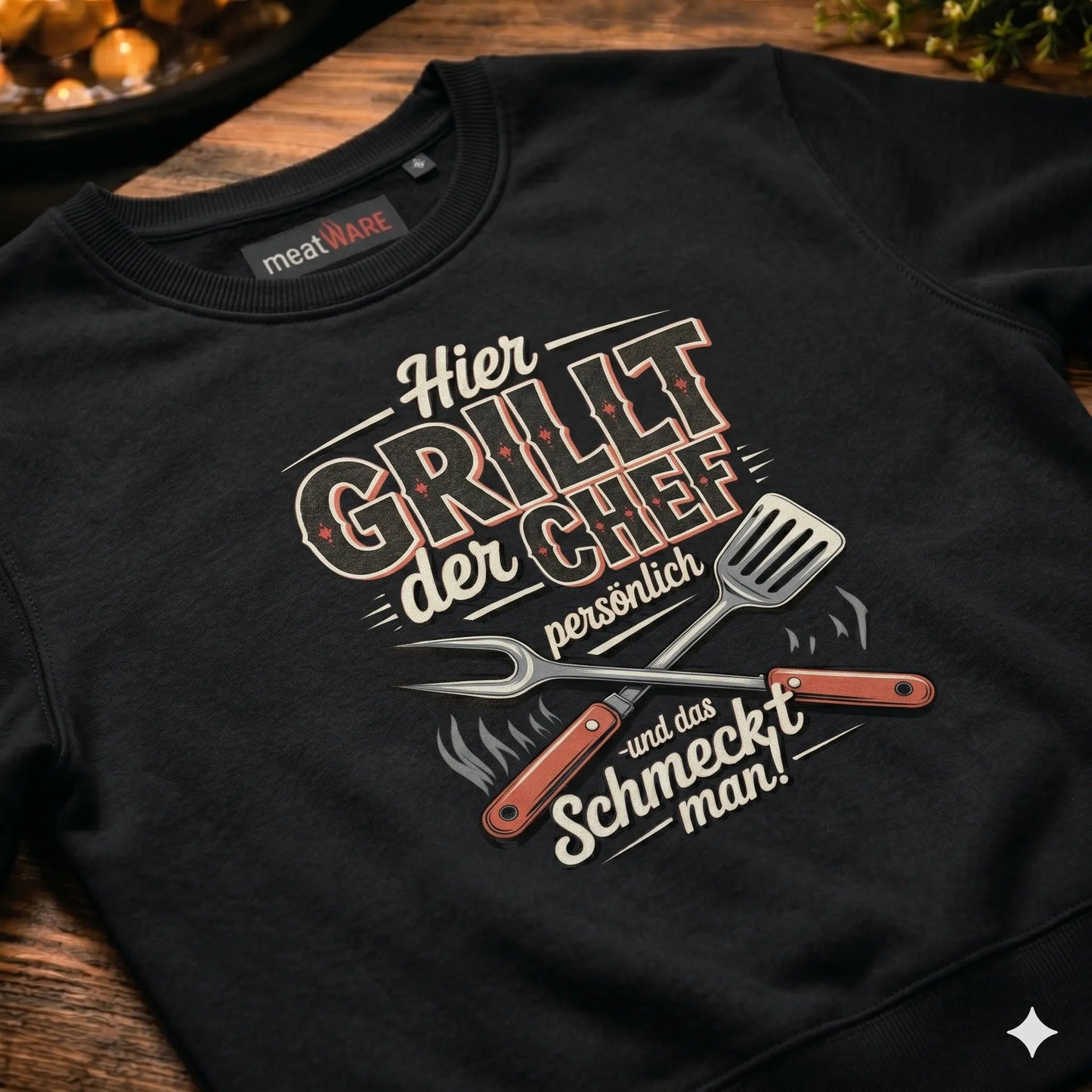 Nahaufnahme eines schwarzen Premium Organic Sweatshirts mit dem "Hier GRILLT der CHEF persönlich"-Grafik und gekreuzten Grillutensilien, präsentiert in einer BBQ-Küchenszene auf einem Holzgrund mit dem "meatWARE"-Nackenlabel.