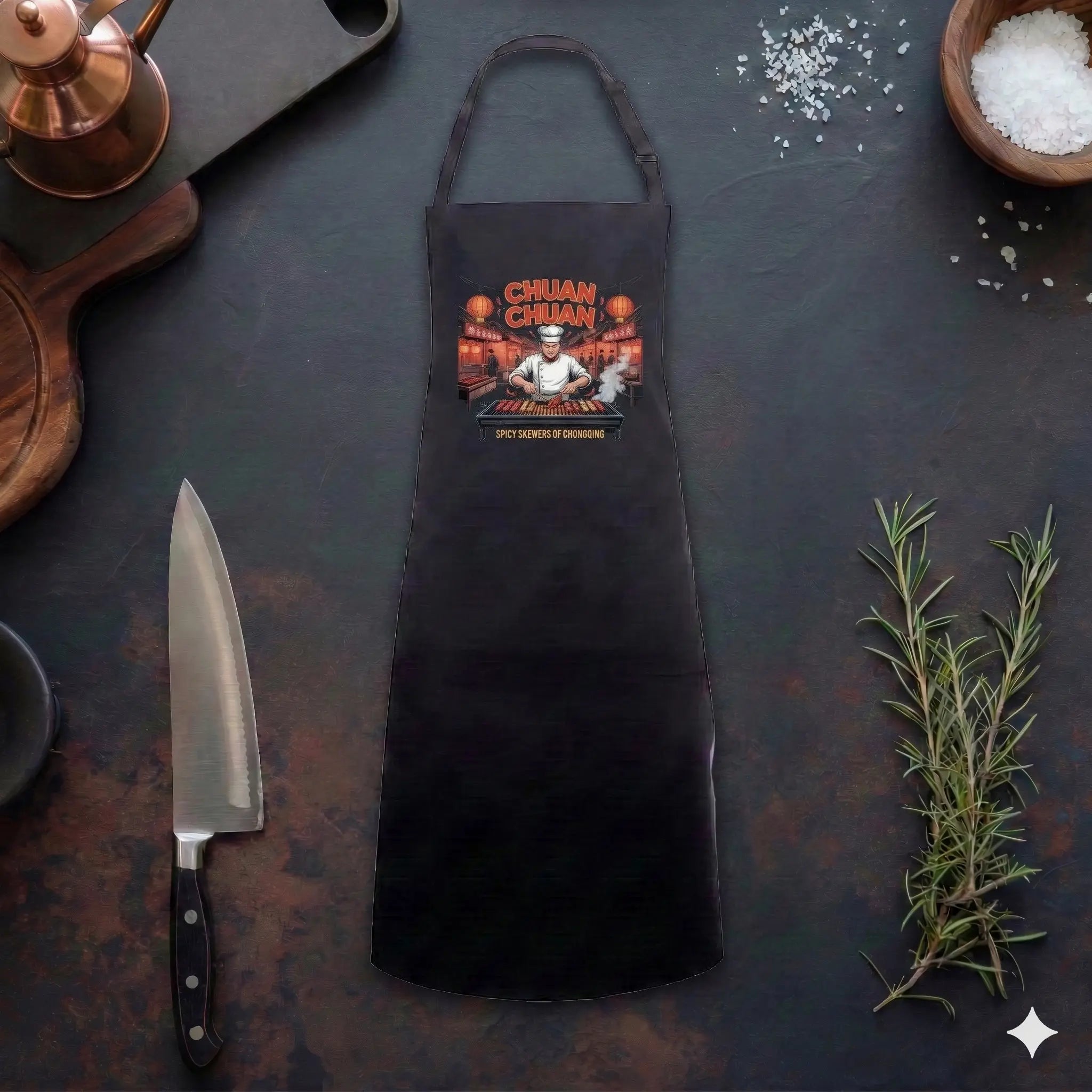 Eine flache Draufsicht auf eine schwarze Chuan Chuan Grillschürze, die auf einer rustikalen dunklen Küchentheke liegt. Das detaillierte Illustrations-Motiv auf der Schürze zeigt einen lächelnden chinesischen Koch mit Kochmütze, der über einem Grill mit vielen Spießen steht, umgeben von einem belebten Nachtmarkt mit roten Laternen und dem Text "CHUAN CHUAN" sowie "SPICY SKEWERS OF CHONGQING". Die Schürze ist umgeben von Kochutensilien wie einem großen Kochmesser, einem hölzernen Schneidebrett, einem kupferne