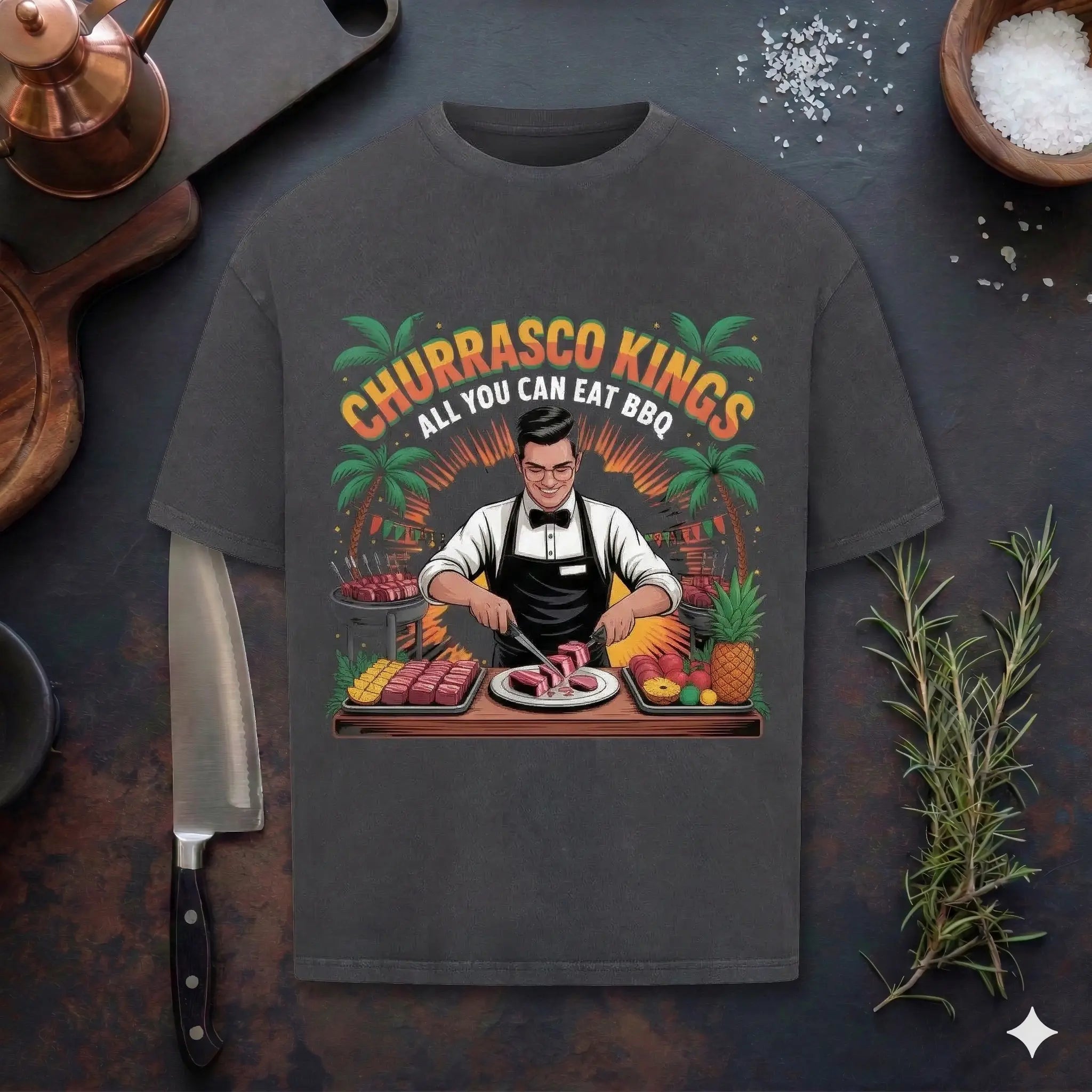 Eine flache Draufsicht auf ein schwarzes, übergroßes T-Shirt aus schwerer Baumwolle auf einer rustikalen Küchentheke. Das T-Shirt zeigt eine detaillierte Vintage-Illustration eines lächelnden brasilianischen Churrasco-Kellners mit Fliege und Schürze, der Picanha-Fleisch am Tisch schnitzt. Über der Illustration steht in großer, stilisierter Schrift "CHURRASCO KINGS" in Gelb und Grün, darunter "ALL YOU CAN EAT BBQ". Der Kellner ist umgeben von Fleischspießen, Ananas, anderen Früchten, einem Grill und Palmen i