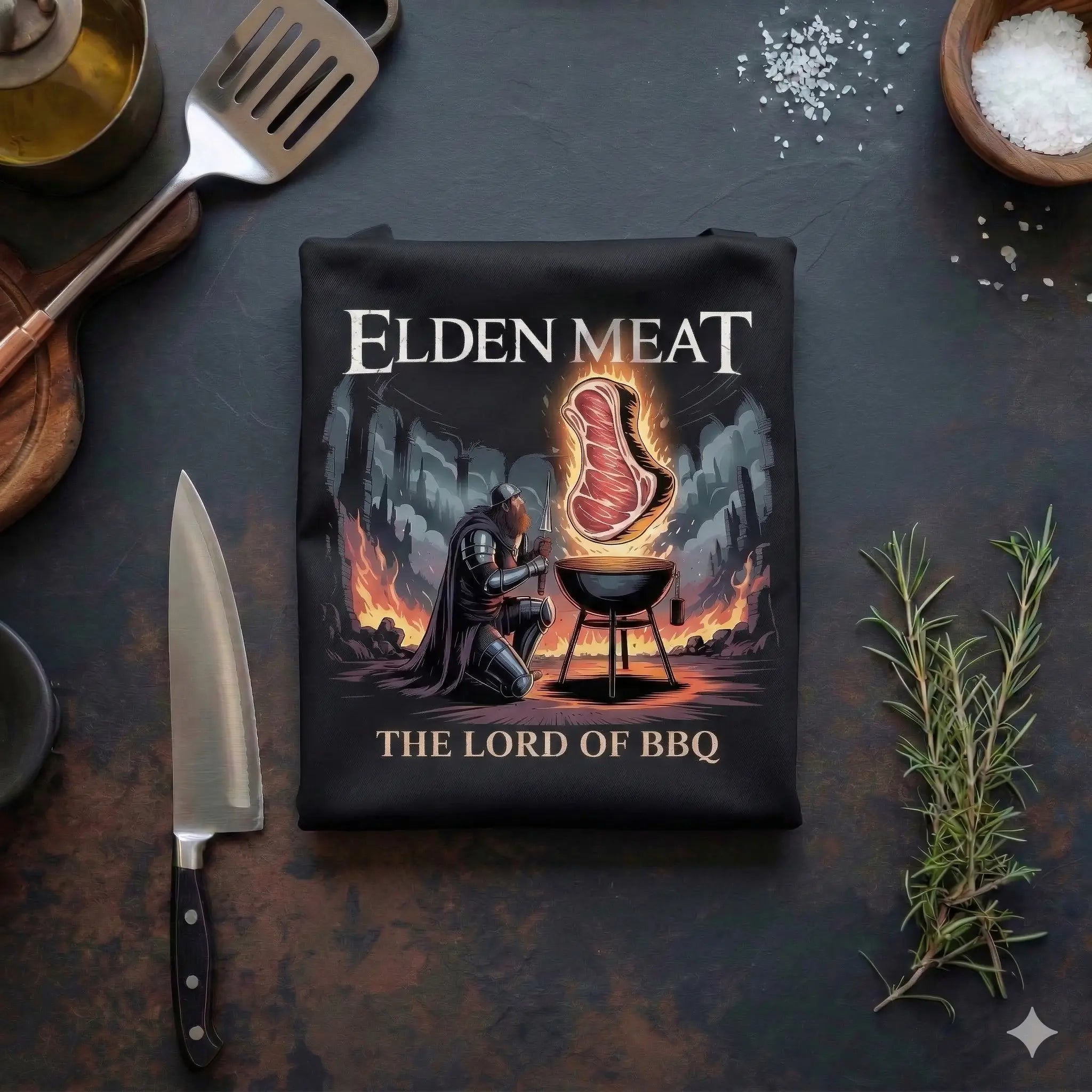 Eine schwarz gefaltete Grillschürze mit dem Aufdruck "Elden Meat - The Lord of BBQ", die flach auf einer dunklen Arbeitsplatte neben Grillutensilien wie einem Messer, einem Pfannenwender und frischem Rosmarin liegt
