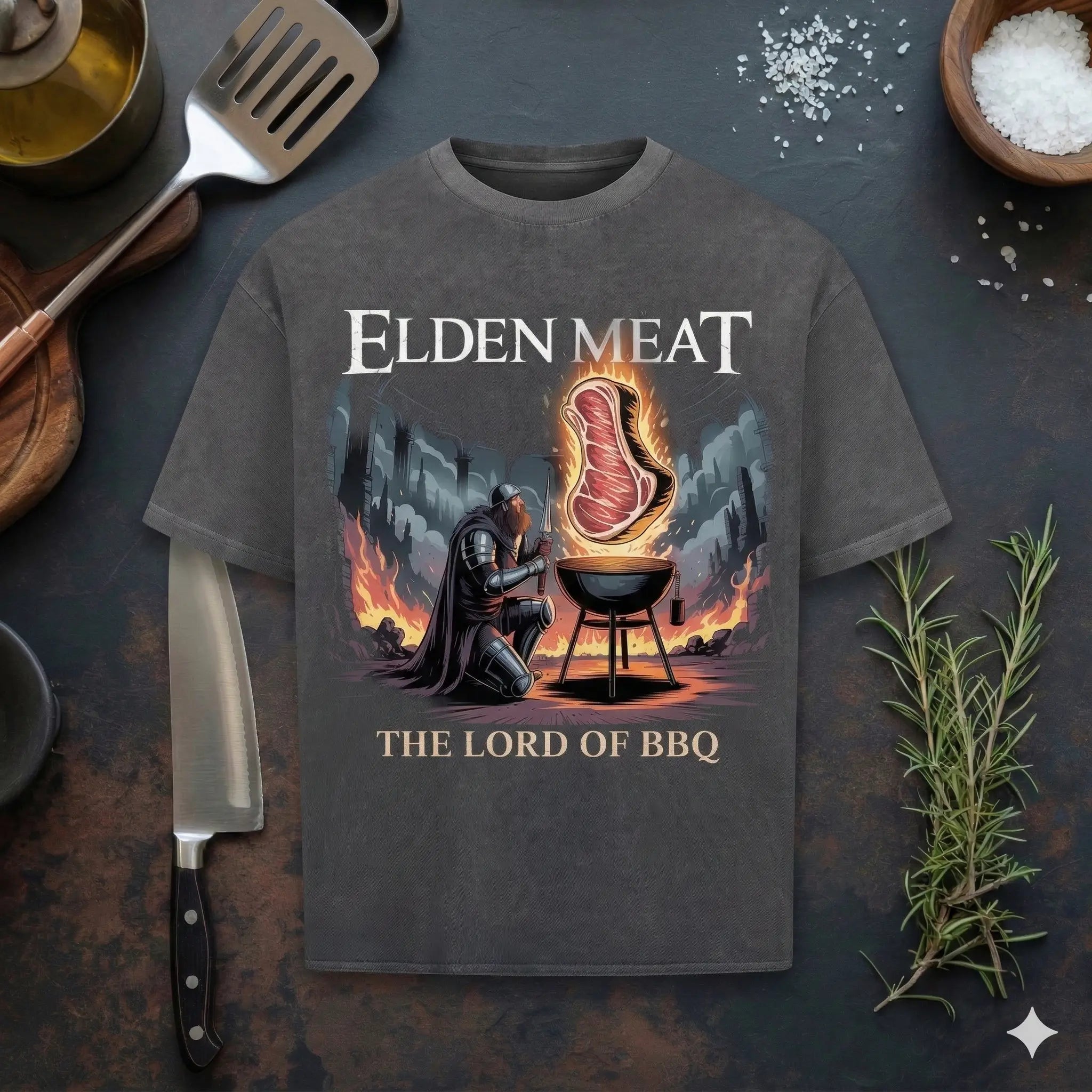 Ein dunkelgraues, gewaschenes Oversized-T-Shirt mit dem Grafikdesign „Elden Meat – The Lord of BBQ“, flach liegend auf einem dunklen Holztisch neben Küchenutensilien wie einem Messer, Rosmarin und einer Schale Salz. Das Design zeigt einen Ritter in Rüstung, der vor einem Grill kniet, über dem ein brennendes Steak schwebt.