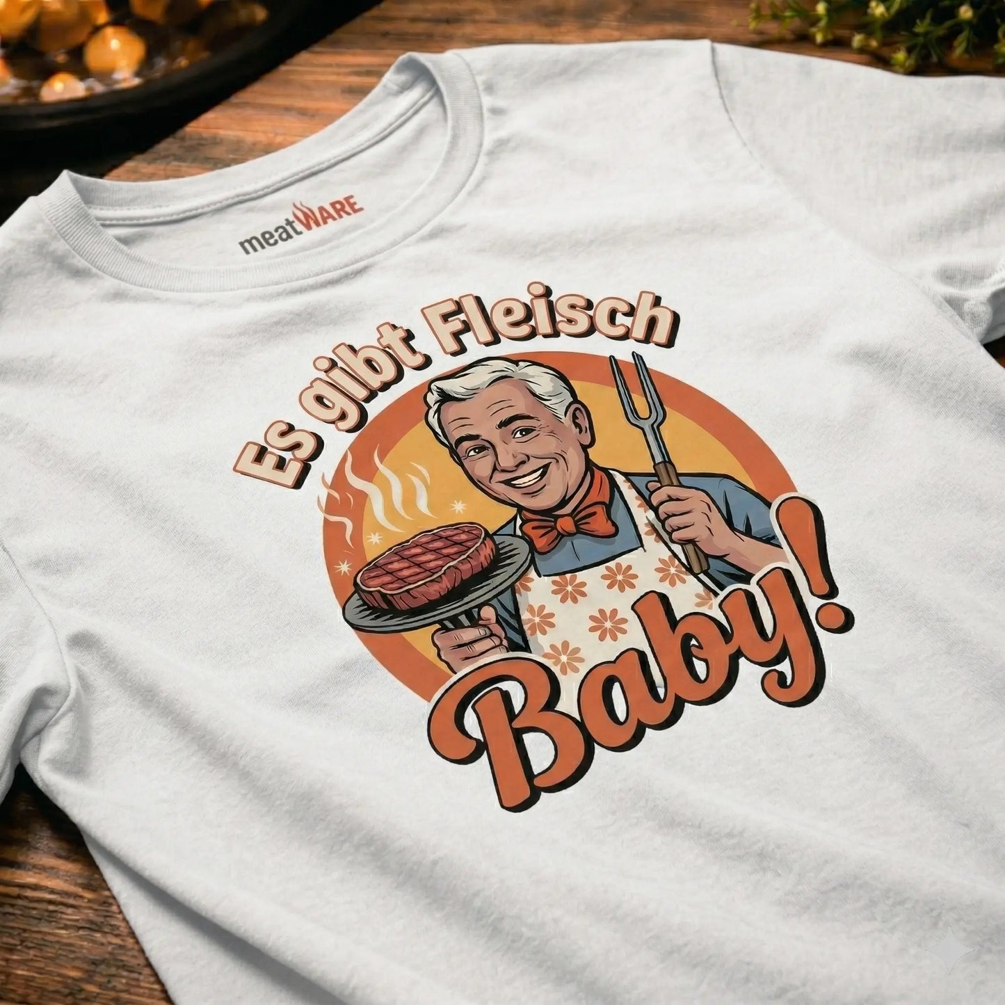 Ein weißes T-Shirt im Vintage-Stil, das flach auf einer Holzoberfläche liegt. Der deutsche Aufdruck zeigt einen lächelnden Koch, der einen Grillteller mit einem dampfenden Steak und eine Grillgabel hält. Der Text lautet: 'Es gibt Fleisch, Baby!'.