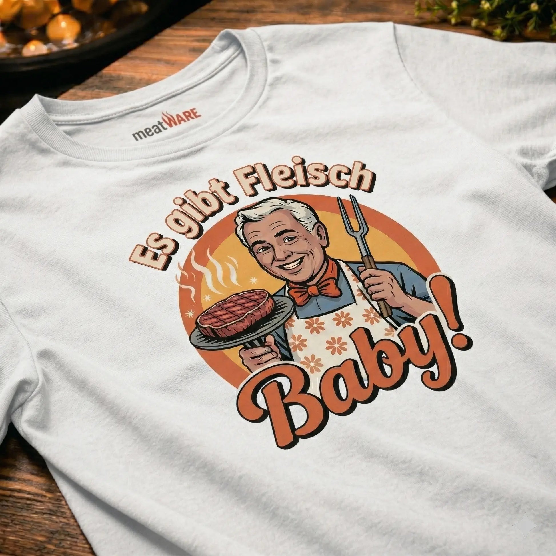 Ein weißes T-Shirt im Vintage-Stil, das flach auf einer Holzoberfläche liegt. Der deutsche Aufdruck zeigt einen lächelnden Koch, der einen Grillteller mit einem dampfenden Steak und eine Grillgabel hält. Der Text lautet: 'Es gibt Fleisch, Baby!'.