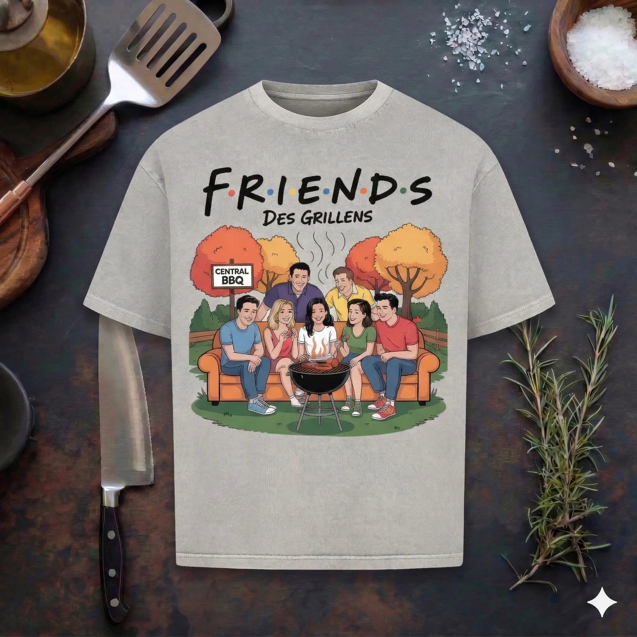 Ein ashgraues T-Shirt mit Melange-Optik auf einem dunklen Holzhintergrund. Das Motiv zeigt eine parodistische Illustration der "Friends"-Charaktere beim gemeinsamen Grillen im Park unter herbstlichen Bäumen, ergänzt durch den Schriftzug "FRIENDS DES GRILLENS" und ein "Central BBQ" Schild.