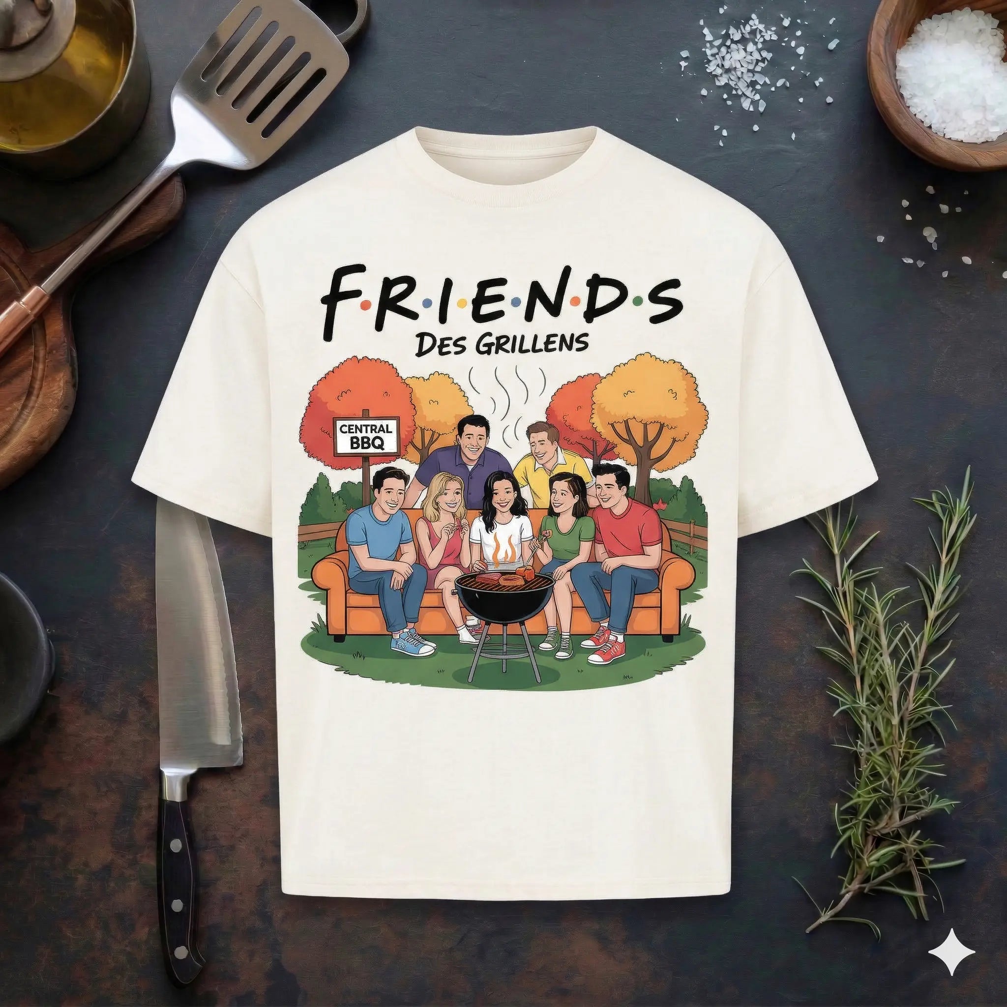 Ein cremefarbenes T-Shirt auf einem dunklen Holzhintergrund mit Grillutensilien. Das Design zeigt eine Comic-Illustration der "Friends"-Charaktere an einem Grill im Freien mit dem Schriftzug "FRIENDS DES GRILLENS" und einem "Central BBQ" Schild.