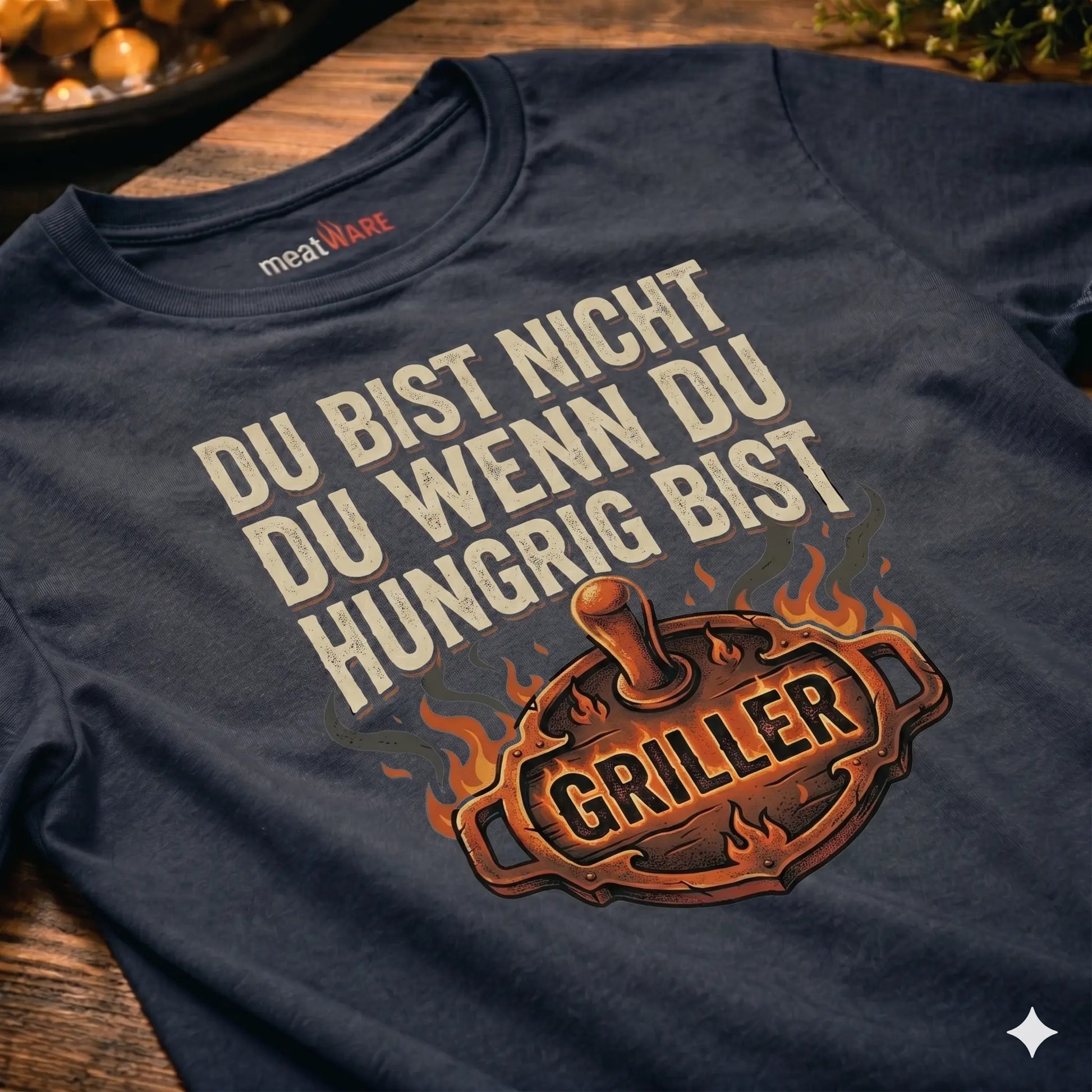 Eine schräge Nahaufnahme eines dunkelgrauen Oversized-T-Shirts im Vintage-Look auf einem rustikalen Holztisch. Das Shirt zeigt einen detaillierten Aufdruck mit einem flammenden Grillgerät und dem Text "GRILLER". Darüber steht in cremefarbener Schrift: "DU BIST NICHT DU WENN DU HUNGRIG BIST". Im Nacken ist ein "meatWARE" Etikett sichtbar.