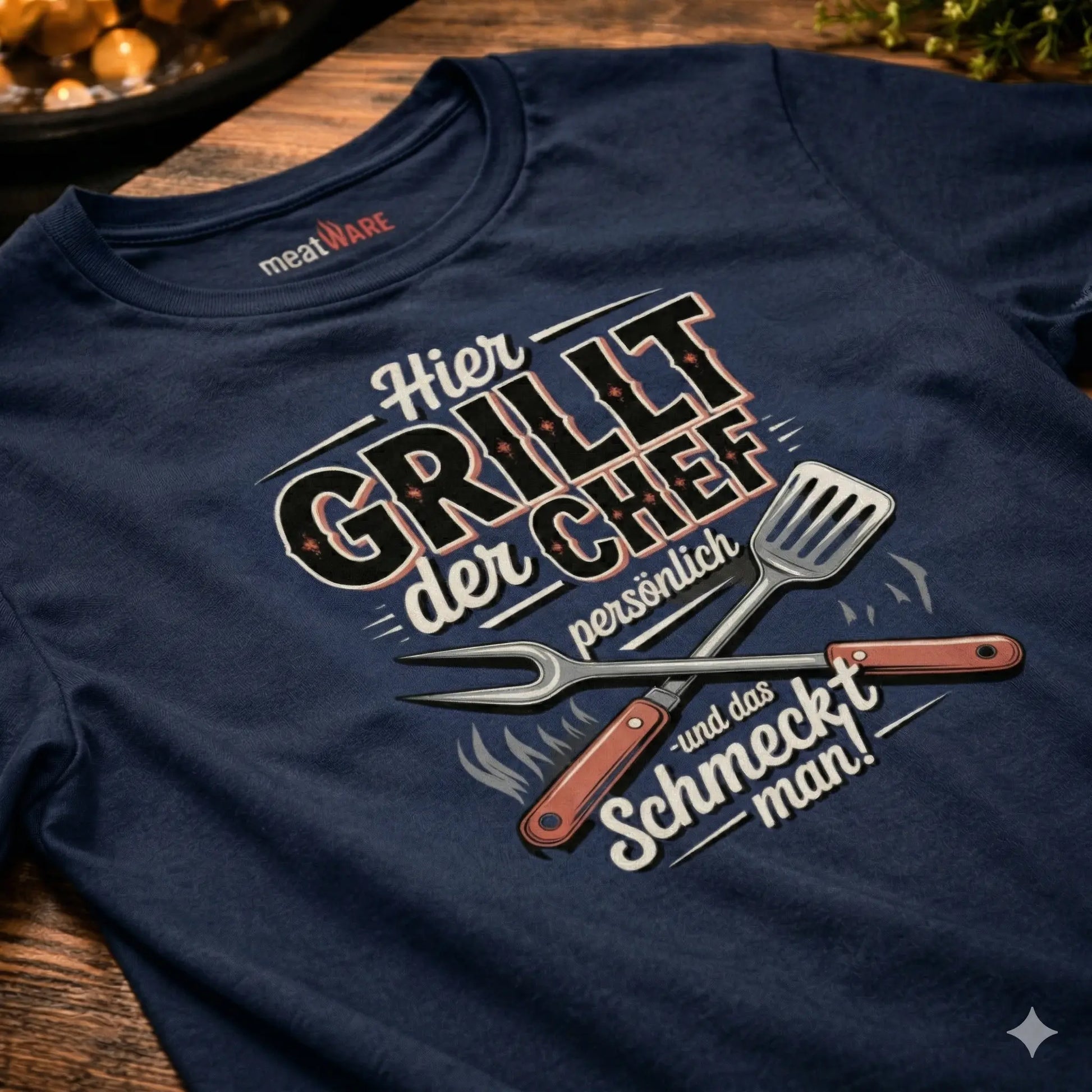 Ein navy-blaues T-Shirt im Vintage-Stil, das auf einer rustikalen Holzoberfläche liegt. Der deutsche Aufdruck zeigt gekreuztes Grillbesteck und den Text: 'Hier grillt der Chef persönlich - und das schmeckt man!'.