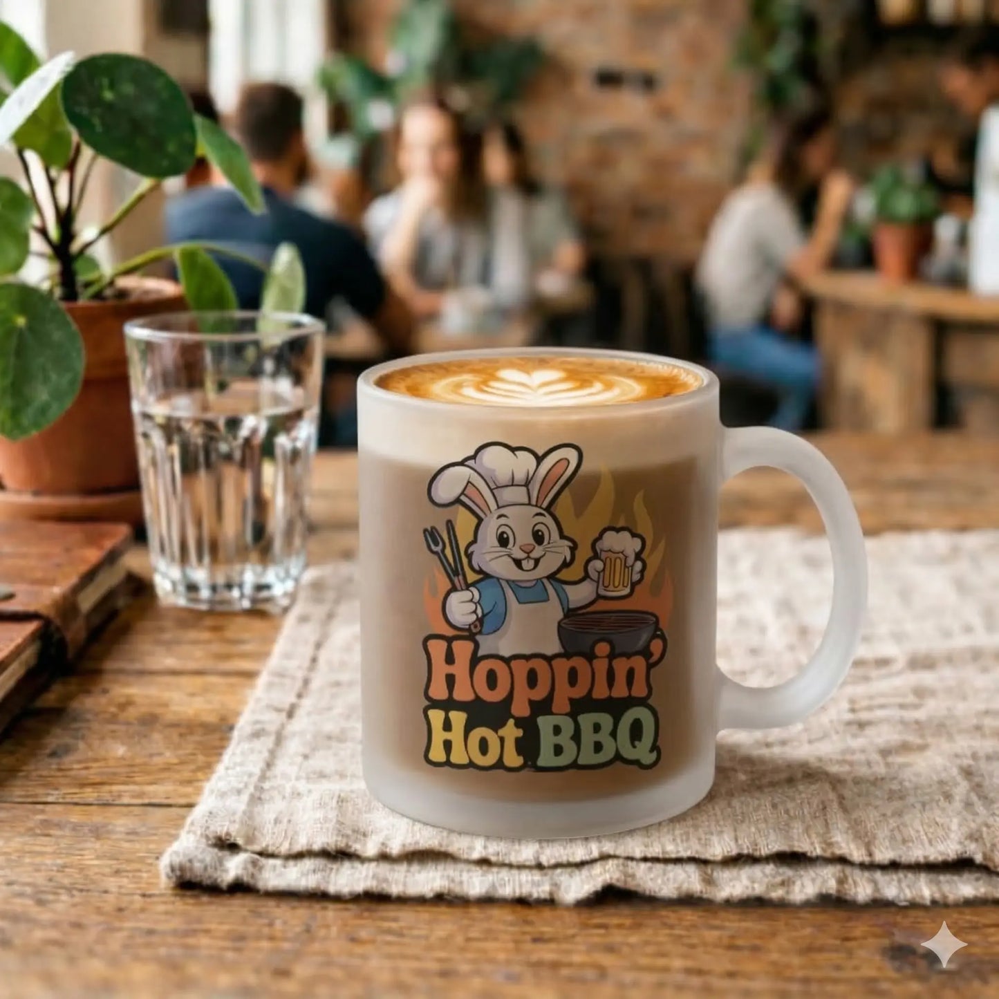 Eine satinierte Glastasse mit dem Aufdruck eines Hasenkochs und dem Text "Hoppin' Hot BBQ" steht auf einem rustikalen Holztisch in einem belebten Café. Die Tasse ist mit einem Cappuccino mit Latte Art gefüllt. Im Hintergrund sind Pflanzen und Gäste zu sehen.