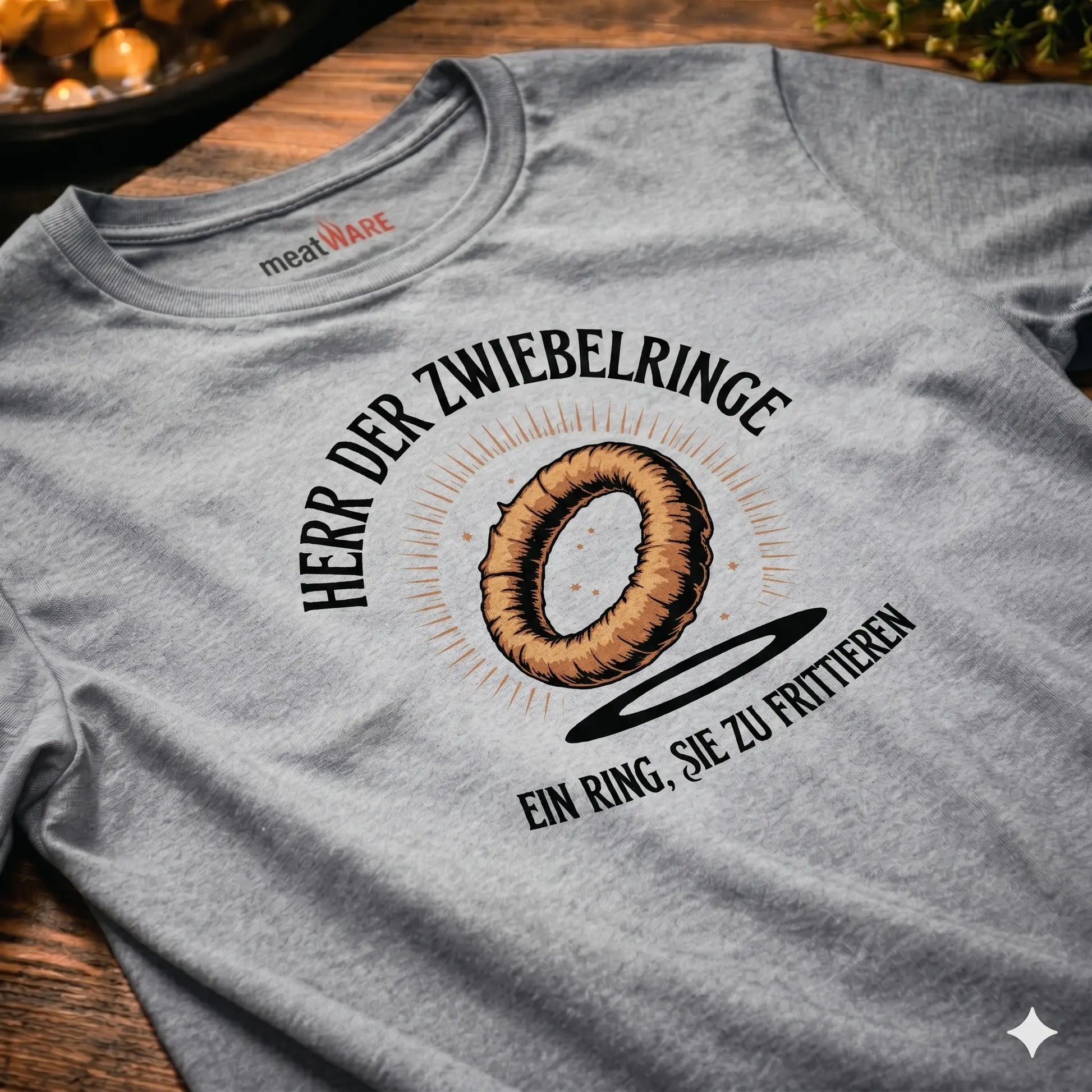 Nahaufnahme eines schräg auf einem rustikalen Holztisch liegenden, hellgrau melierten Premium-T-Shirts mit dem „Herr der Zwiebelringe“-Design. Das Design zeigt einen schwebenden Zwiebelring mit Heiligenschein und deutschem Text: „Herr der Zwiebelringe“ oben im Bogen und „Ein Ring, sie zu frittieren“ darunter. Das T-Shirt hat ein „meatWARE“-Nackenlabel. Die Szene ist warm beleuchtet, mit Unschärfe-Lichtern im Hintergrund und grünem Blattwerk oben rechts.