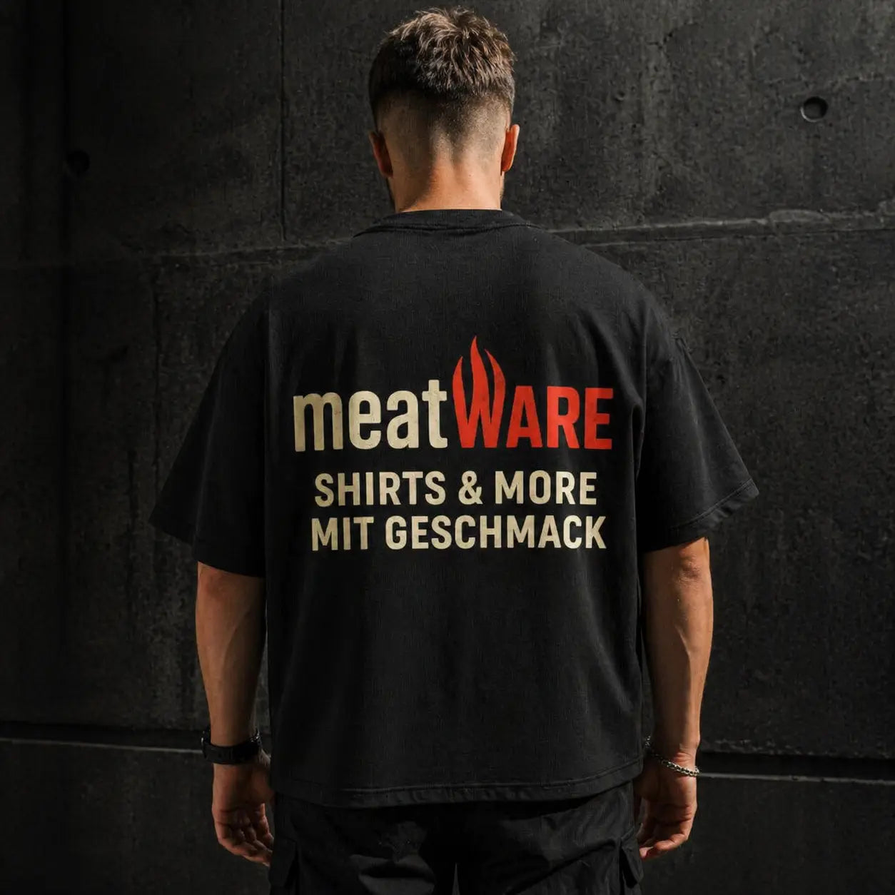Oversized schwarzes Meatware T-Shirt mit großem Backprint „Shirts & More mit Geschmack“, getragen vor dunkler Betonwand, Streetwear Look mit lockerem Schnitt und starkem Markenstatement im Fokus
