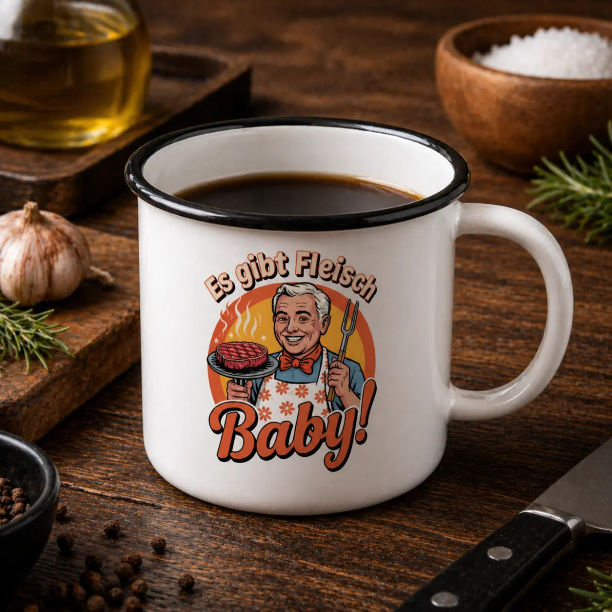 Weiße Emaille Tasse mit schwarzem Rand und „Es gibt Fleisch Baby“ Grill Design, gefüllt mit Kaffee auf rustikalem Holztisch mit Gewürzen und Küchenutensilien in Nahaufnahme