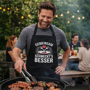 Model beim Grillen trägt schwarze Meatware Grill- und Kochschürze mit Motiv „Gemeinsam schmeckt’s besser“ (Teller, gekreuzte Gabeln, Herz) – Love goes through the stomach.