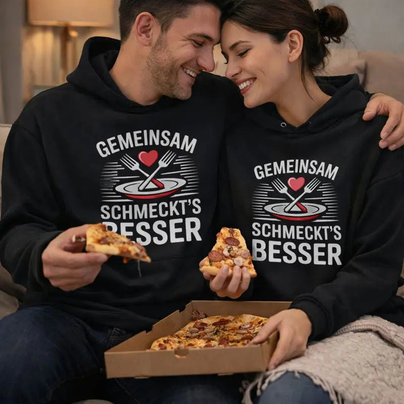 Glückliches Paar auf der Couch teilt Pizza und trägt schwarze Meatware Hoodies mit dem Spruch „Gemeinsam schmeckt’s besser“ – Liebe geht durch den Magen, gemütlicher Abendlook.