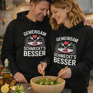 Verliebtes Paar trägt schwarze Meatware Hoodies mit dem Statement „Gemeinsam schmeckt’s besser“ und genießt Wein und Brotzeit bei stimmungsvoller Beleuchtung – Liebe geht durch den Magen.