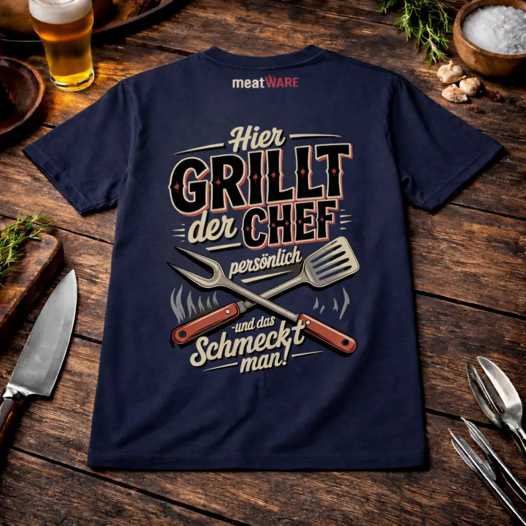 Navy blaues Grill T-Shirt mit Aufdruck „Hier grillt der Chef persönlich und das schmeckt man“ im rustikalen BBQ Mockup mit Bier, Knoblauch und Grillbesteck auf Holztisch sowie Meatware Logo im Nackenbereich