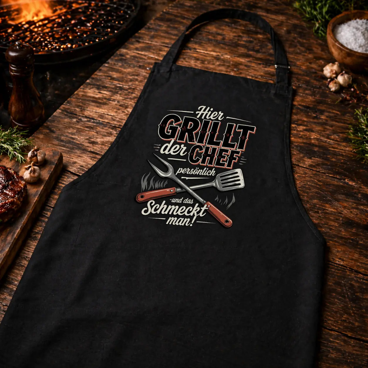 Schräg fotografierte Nahaufnahme einer schwarzen Meatware Grillschürze mit Motiv „Hier grillt der Chef persönlich und das schmeckt man“ im rustikalen BBQ Mockup auf Holztisch mit Grill und Zubehör