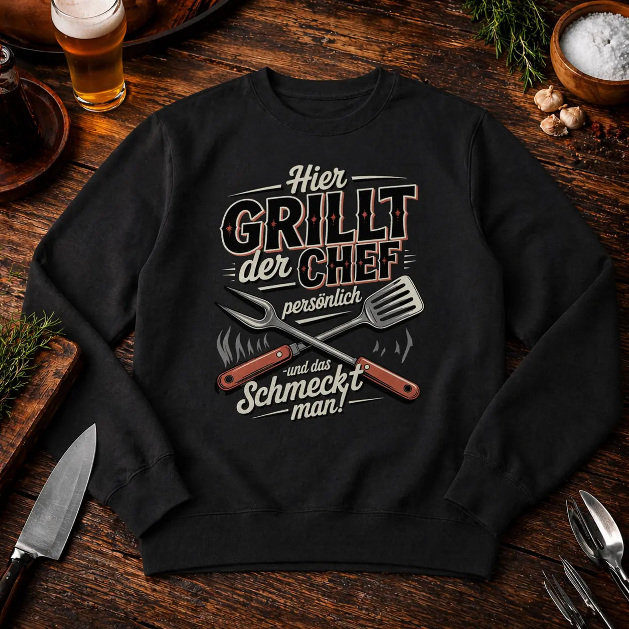 Schwarzes Premium Organic Sweatshirt mit „Hier grillt der Chef persönlich“ BBQ Motiv im rustikalen Grill Mockup auf Holztisch mit Bier und Grillzubehör