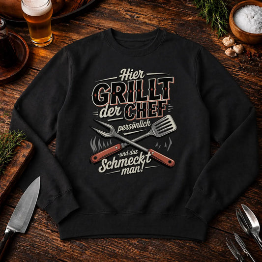 Schwarzes Premium Organic Sweatshirt mit „Hier grillt der Chef persönlich“ BBQ Motiv im rustikalen Grill Mockup auf Holztisch mit Bier und Grillzubehör