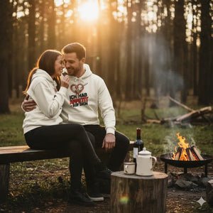 Paar am Lagerfeuer im Wald, trägt cremefarbene Hoodies mit Herz-Nudeln-Print, Sonnenuntergang, warme Atmosphäre.