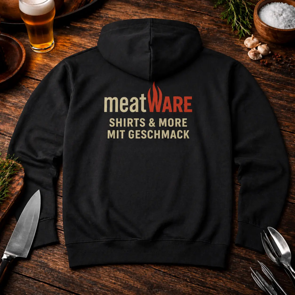 Schwarzer Meatware Premium Hoodie mit Frontlogo auf rustikalem Holztisch, umgeben von Grillbesteck, Bier und Gewürzen in BBQ Atmosphäre