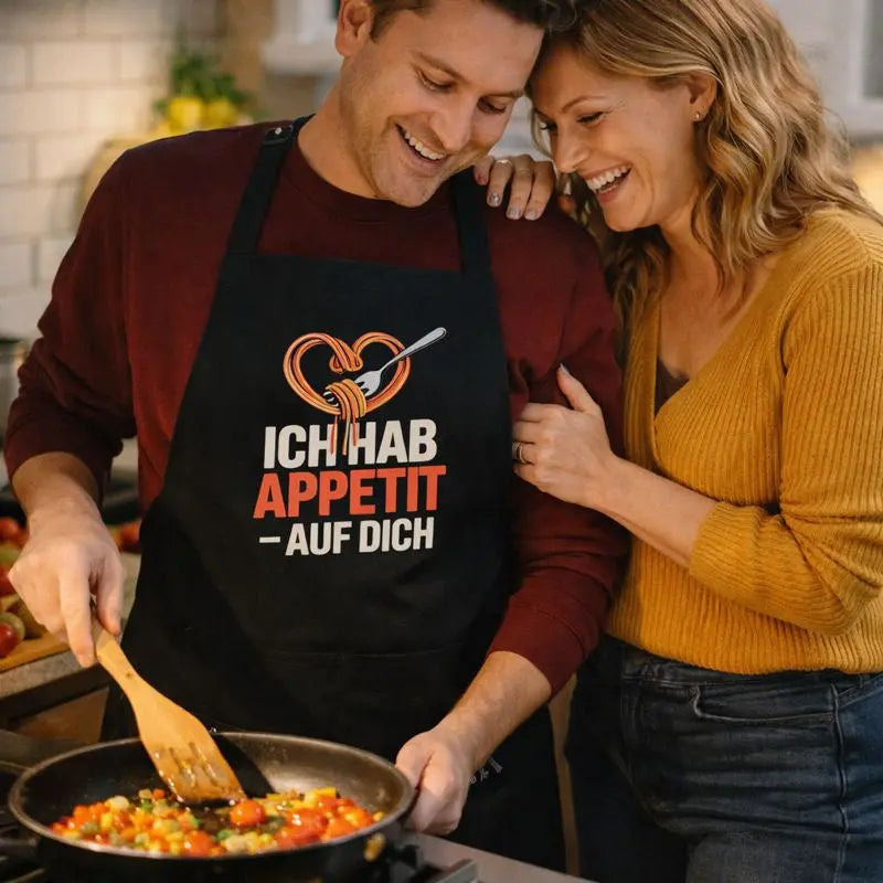 Paar kocht gemeinsam und trägt schwarze Meatware Grill- und Kochschürze mit Spaghetti-Herz-Motiv und Schriftzug „Ich hab Appetit auf Dich“ – humorvolles Liebesdesign zum Thema Liebe geht durch den Magen