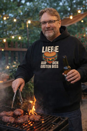 Mann mit Bauch grillt im Garten und trägt Meatware Oversized Hoodie „Liebe auf den ersten Biss“ beim BBQ am Abend.