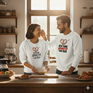 Lachendes Paar in weißen Organic Sweatshirts beim gemeinsamen Backen in einer lichtdurchfluteten, rustikalen Küche.