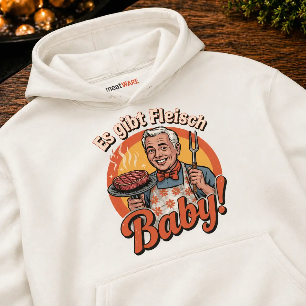 Oversized Hoodie in Cream mit Retro Grill Design „Es gibt Fleisch Baby“ in Nahaufnahme auf Holztisch, Meatware Hoodie mit Steak Motiv und Grillmeister Illustration