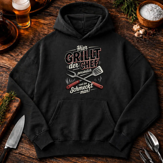 Schwarzer Oversized Hoodie im BBQ Flatlay Mockup auf rustikalem Holztisch mit Bier, Gewürzen und Grillbesteck, großes Druckmotiv zentral platziert