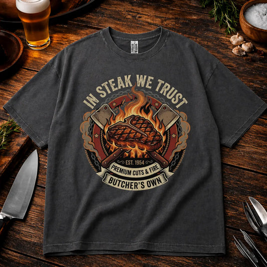 Graphitfarbenes Oversized T-Shirt mit „In Steak We Trust“ Grillmotiv im rustikalen BBQ Mockup auf Holztisch mit Bier, Messer und Gewürzen, lockerer Schnitt, realistische Stoffstruktur und hochwertiger Druck.