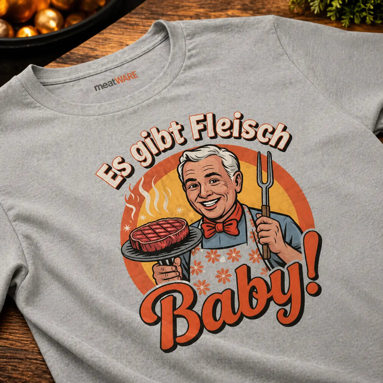 Oversized T-Shirt in Grau mit Retro Grill Design „Es gibt Fleisch Baby“ in Nahaufnahme auf Holztisch, Meatware Shirt mit Steak Motiv und Grillmeister Illustration